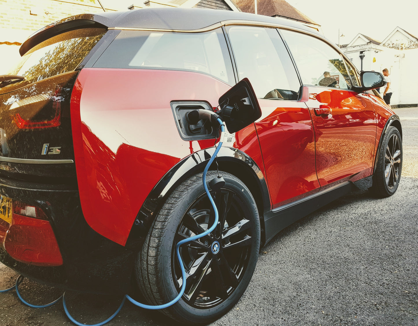 BMW i3s on Charge .  Blue cable being used to charge a red car on gravel with a building in the background